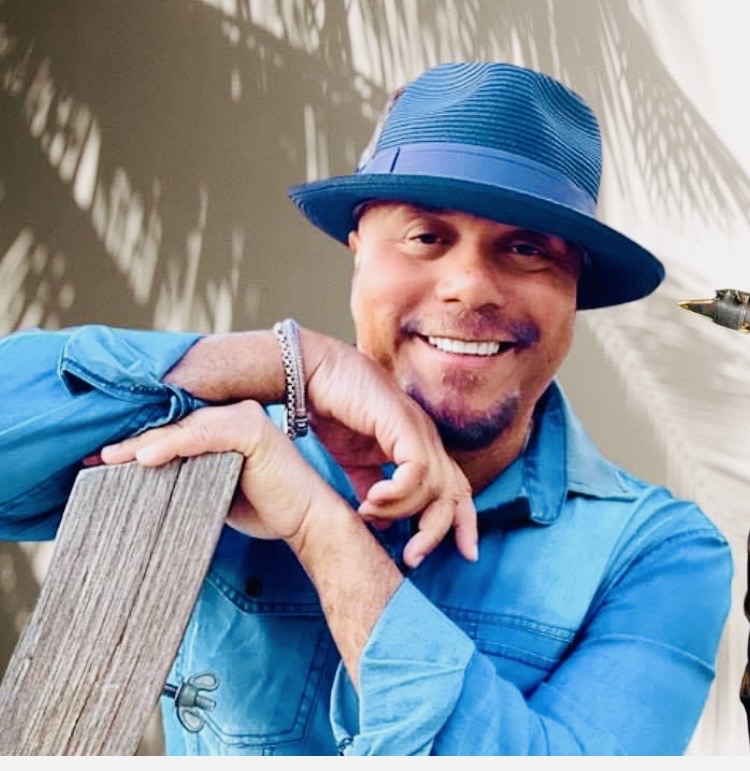 Man smiling in a blue shirt and hat, leaning on a wooden post.