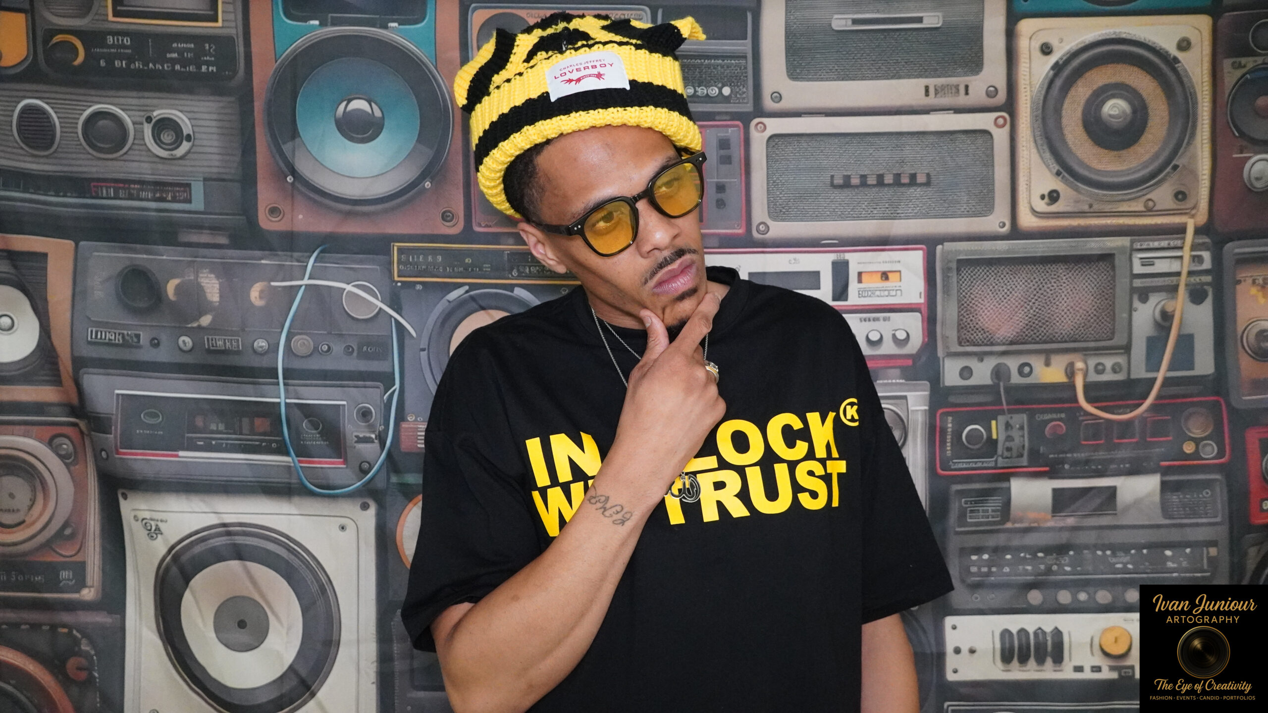 Man in yellow-striped hat and sunglasses posing thoughtfully against a wall of vintage speakers.