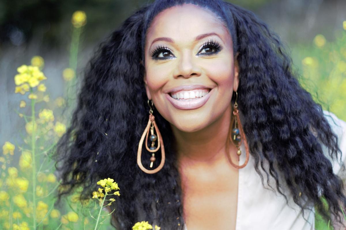 Smiling woman with long curly hair and large earrings in a natural setting.